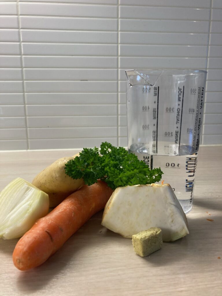 Kuvassa on keittötasolla sipuli, peruna, porkkana, persilja, selleri, kasvisliemikuutio ja vettä mitta-astiassa.